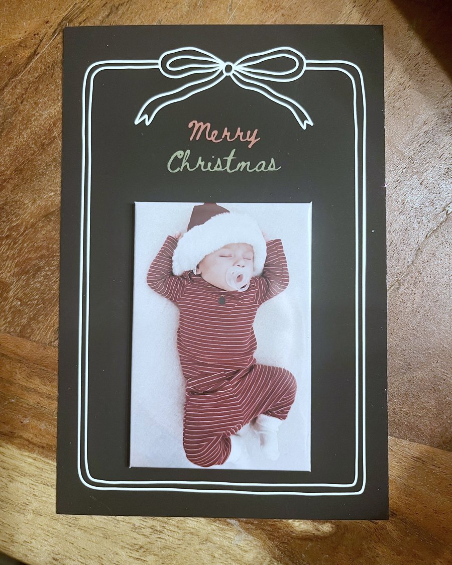 A black chalkboard-style Merry Christmas keepsake card with a white bow and a photo magnet of the same sleeping baby